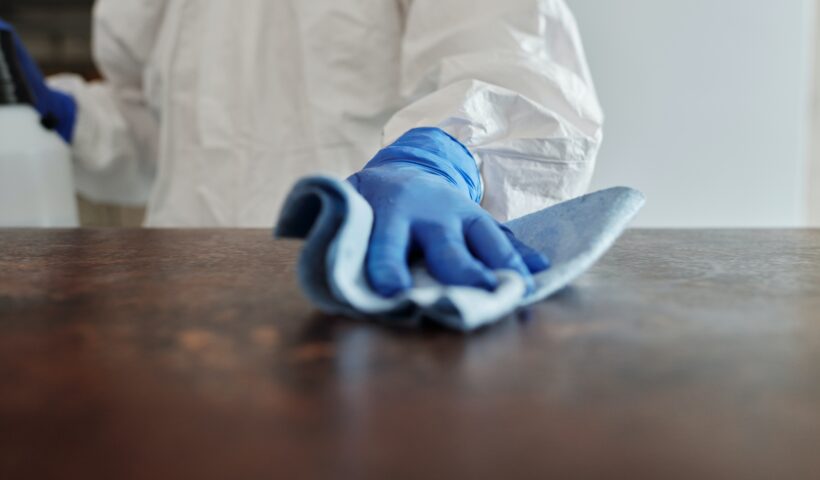 Person in cleaning suit with gloves on wiping down a table.